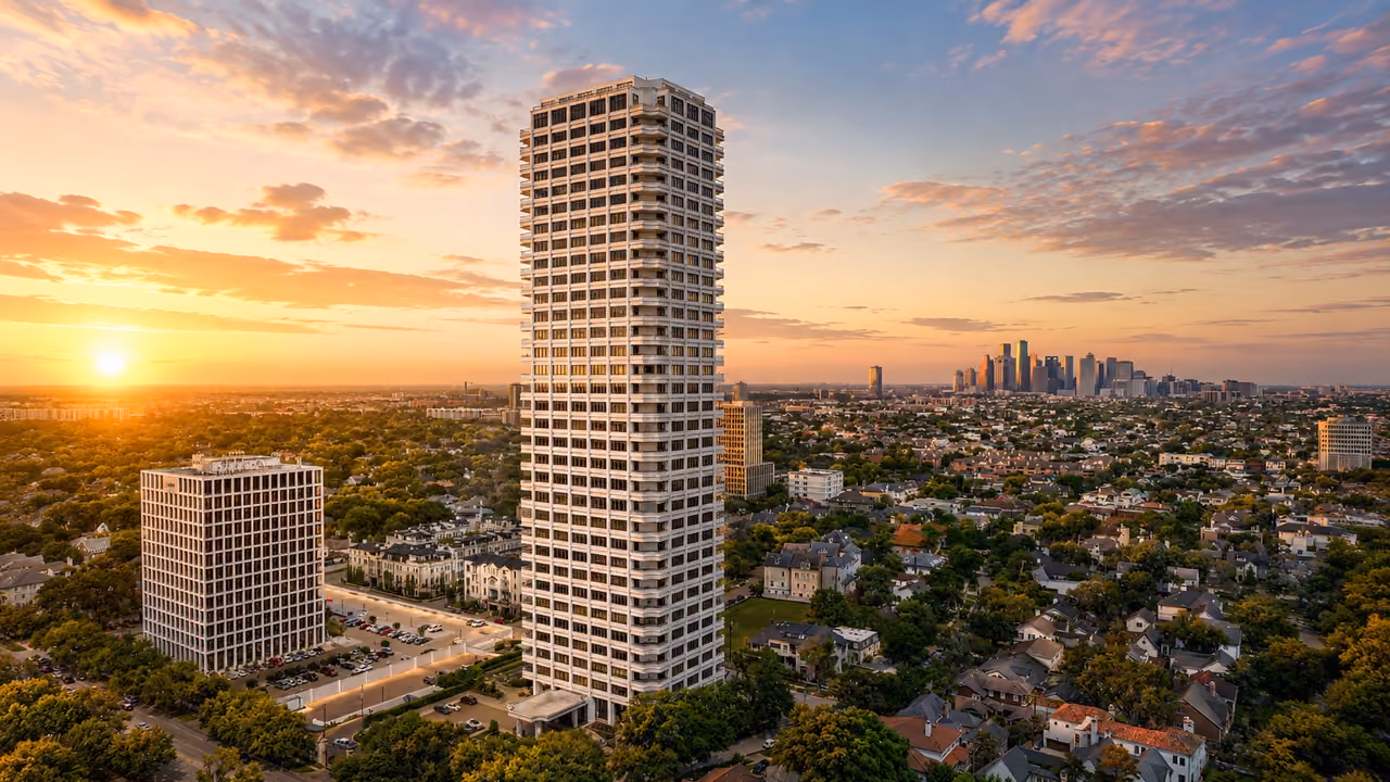 Aerial photograph of The Huntingdon at 2121 Kirby Drive in low golden light: a pale faceted tower banded with recessed balconies stands alone at the centre of the frame, rising well above the tree-covered residential blocks around it, its west face lit gold by a sun low at the far left. A lower cream office building with a grid of square windows and a surface car park sits to the left, a row of white townhouses and a smaller tan mid-rise stand behind the tower, and pitched-roof houses under mature trees fill the right of the frame and the foreground. The downtown Houston skyline sits on the horizon to the right under gold and lavender clouds.